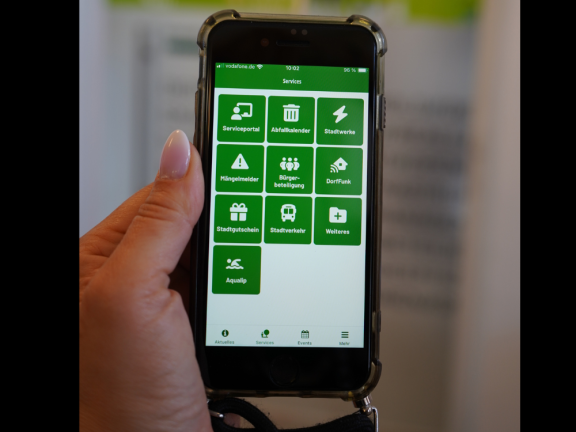 Das Foto zeigt ein in die Höhe gehaltenes Smartphone vor einem unscharfen Hintergrund. Auf dem Bildschirm sieht man die Servicekacheln der städtischen App 