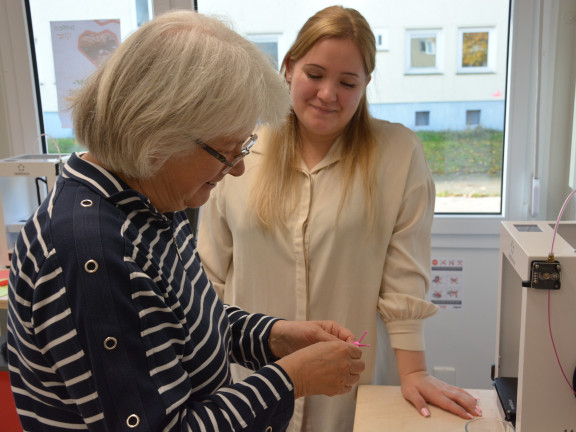 Eine junge Mitarbeiterin der Stadt Detmold erklärt einer Seniorin den 3D-Druck direkt am Gerät. beide Personen stehen. 