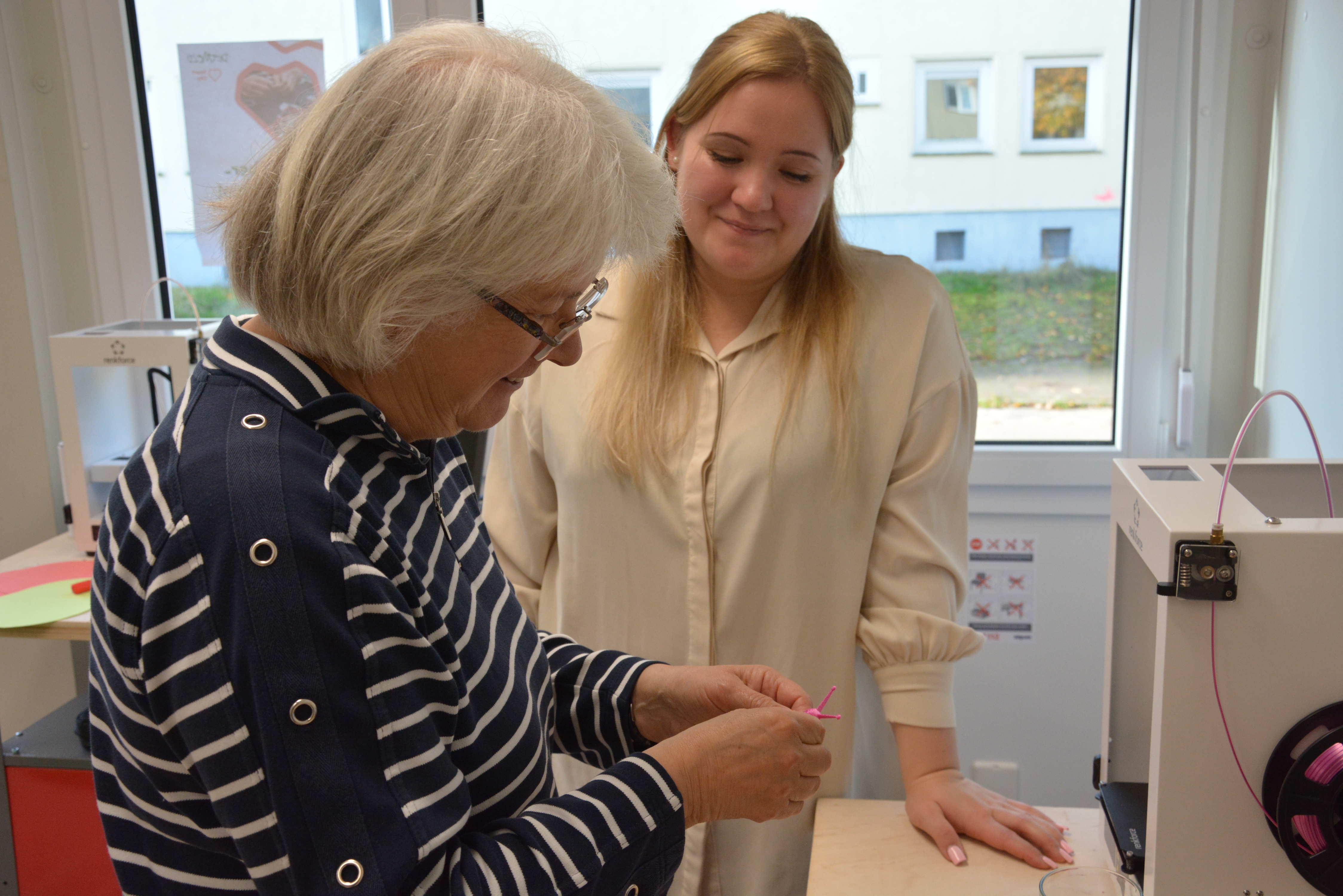 Eine junge Mitarbeiterin der Stadt Detmold erklärt einer Seniorin den 3D-Druck direkt am Gerät. beide Personen stehen.