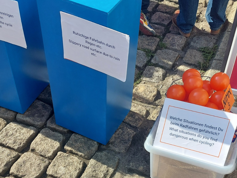 Das Foto zeigt links angeschnitten zwei blaue Abstimmungssäulen aus Holz, die auf Kopfsteinpflaster stehen. An den Säulen kleben Zettel mit Aussagen zum Radverkehr. Rechts steht eine weiße Kiste mit zahlreichen orangenen Bällen, die zur Abstimmung genutzt werden dürfen.