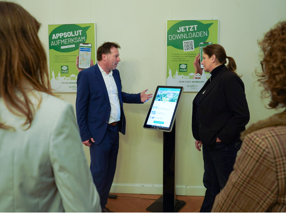 Das Foto zeigt - zwischen Besuchern fotografiert, einen Mann und eine Frau im Gespräch. Sie stehen an einer Infoastele auf der die Stadtapp Appmold läuft. Im Hintergrund sind großflächige, in grün gehaltene Plakate mit Informationen zur Appmold zu sehen. 