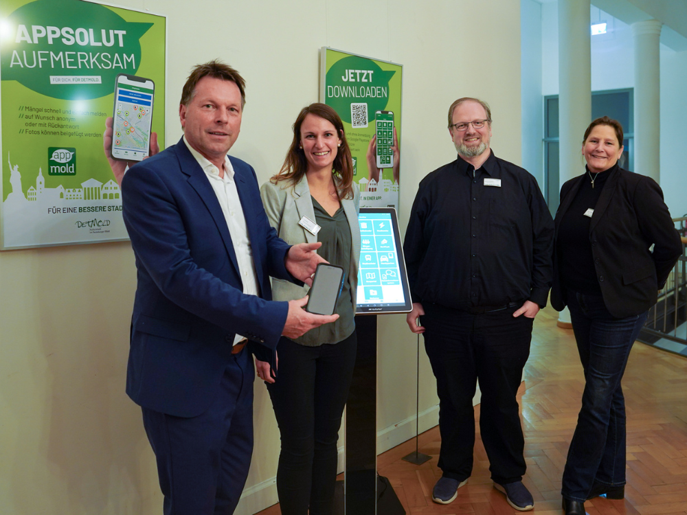 Zwei Männer und zwei Frauen stehen vor der Ausstelleung mit großen grünen Plakaten, die die Stadtapp "Appmold" bewerben. Der Mann links, Bürgermeister Frank Hilker, hält sein Smartphone in die Kamera. 