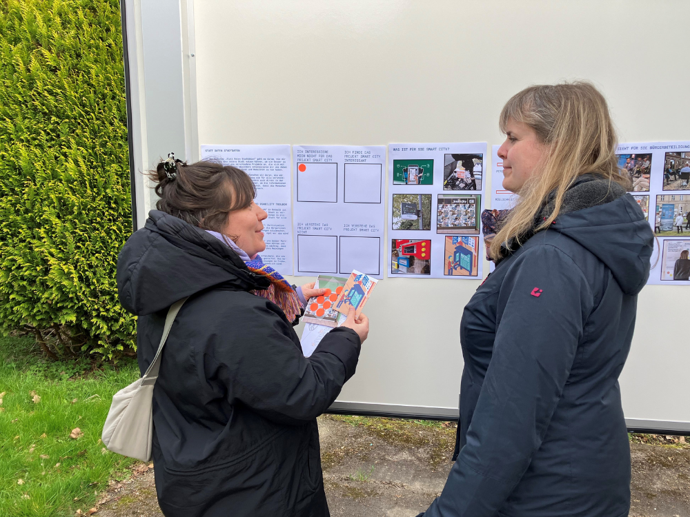Zwei Frauen unterhalten sich vor der Wand des städtischen Anhängers. Im Hintergrund sieht man die Aushänge zum Projekt in DIN A3. Die linke Frau hält rote Klebepunkte in der Hand. 
