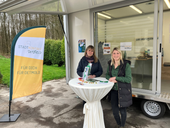 Das Foto zeigt zwei Frauen vor dem städtischen Anhänger. Sie stehen hinter einem Stehtisch mit weißer Husse und lächeln. Links von ihnen steht eine BeachFlag in orange und weiß, auf der 
