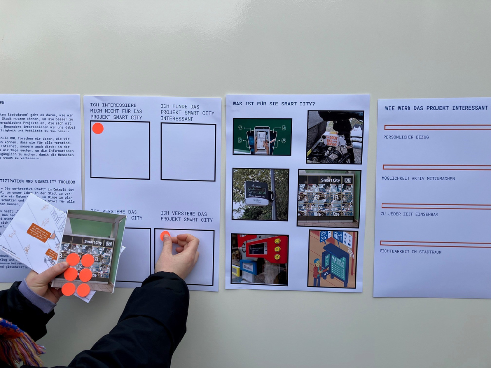 Das Foto zeigt vier Aushänge in DIN A3 Größe. Zwei Hände kleben neonrote Punkte in Kästchen, die eine Abstimmung zu diversen Sachverhalten ermöglichen. Der Text ist so klein, dass man ihn nicht lesen kann. Von der Person sind nur die Hände und Arme zu sehen. 