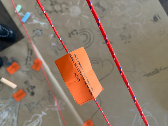 Oranger Zettel mit dem Text 'Alles entwickelt sich, bevor es etwas das sich entfaltet' an rotem Seil befestigt, unscharfer Hintergrund mit Zeichnungen 