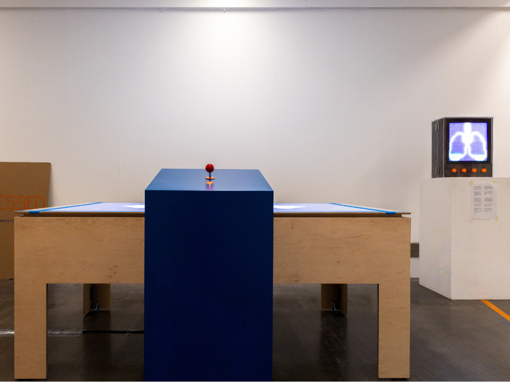 Im Vorderund ist in einem weißen Raum ein großer Schreibtisch aus Holz zu sehen, der als Projektionsfläche für die Visualisierung von Stadtdaten dient. Rechts im Hintergund sieht man eine schwarze Box, auf der eine aus LED-Lichtern fargestellte Lunge dfie Feinstaubbelastung zeigt.