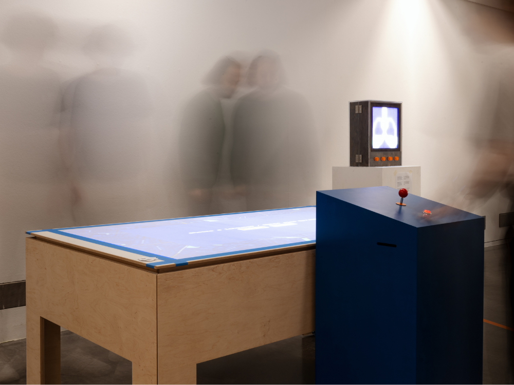 Das Foto zeigt unser zentrales und dezentrales Interface aus dem Smart-City-Projekt. Im Vordergrund steht ein großer Tisch auf den die Daten projiziert werden, rechts sieht man klein das dezentrale Interface, auf dem LED-Leuchten eine Lunge abbilden. Im Hintergrund sind durch die Langzeitbelichtung vier Menschen verschwommen zu erkennen.