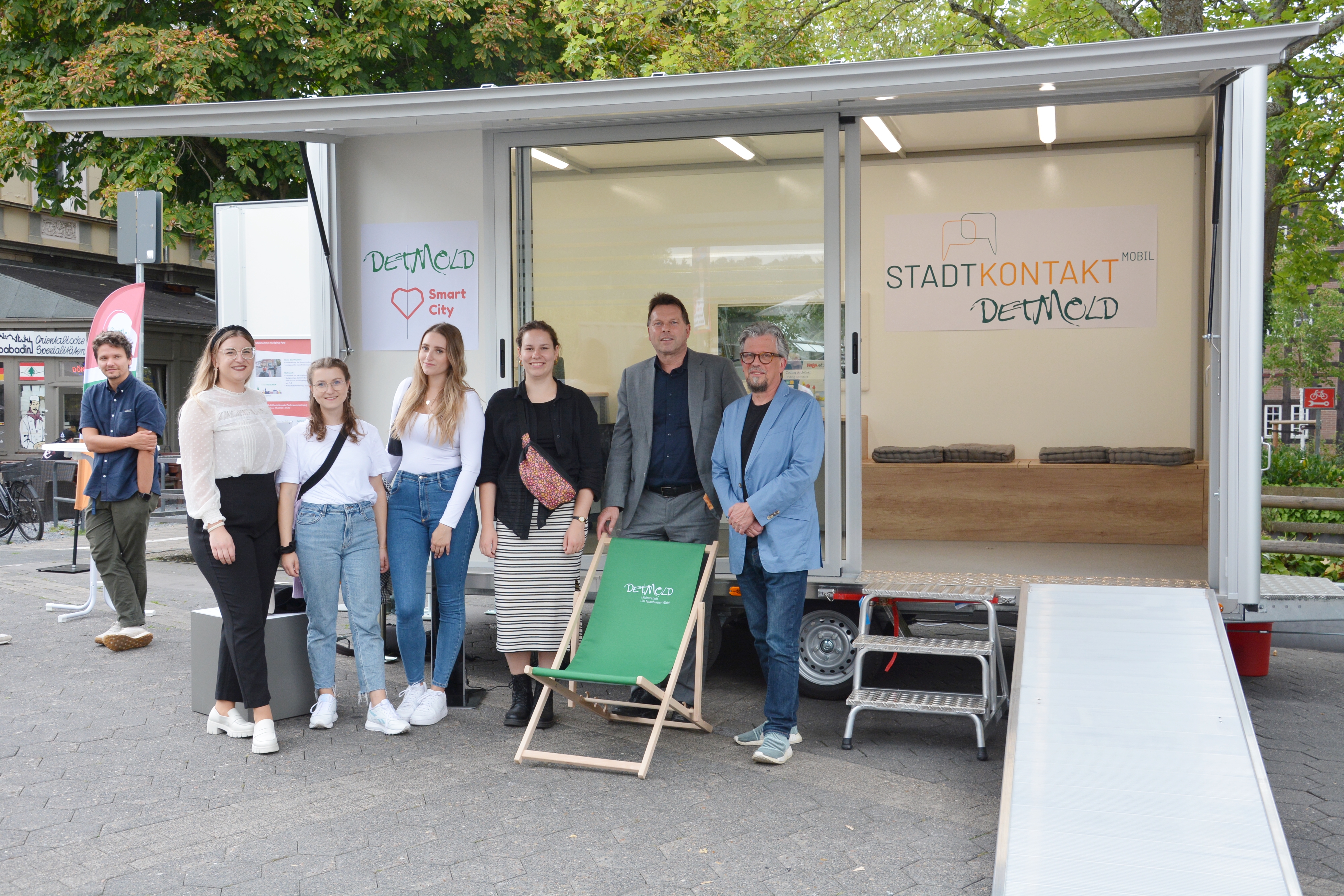 Das Bild zeigt den aufgebauten städtischen Anhänger mit großer Glasfront und ausgeklappter Rampe. Davor steht der Bürgermeister mit einer Gruppe Mitarbeitender der TH OWL.