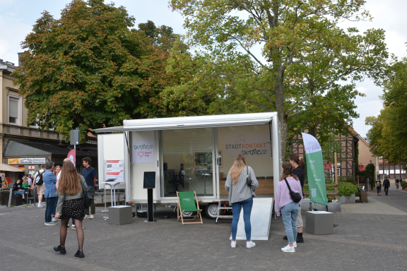 Das Bild zeigt den aufgebauten städtischen Anhänger mit großer Glasfront und ausgeklappter Rampe. Davor stehen Menschen und informieren sich. Diese sind nur von hinten zu sehen. 