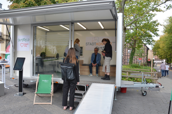 Das Bild zeigt den aufgebauten städtischen Anhänger mit großer Glasfront und ausgeklappter Rampe. Im Anhänger sieht man vier Personen. 