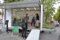 Das Bild zeigt den aufgebauten städtischen Anhänger mit großer Glasfront und ausgeklappter Rampe. Im Anhänger sieht man vier Personen.