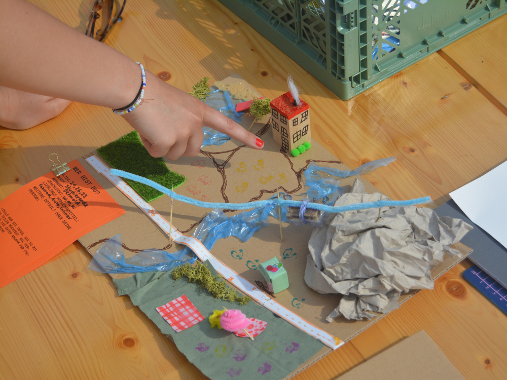 Das Foto eine Aufsicht von oben auf ein gebastelte Stadtmodell, das auf einem Holztisch steht. Eine Hand mit rosa lackierten Nägel zeigt auf das Stadtmodell, das aus Pappe, Stoffen, Moos und Plastikmüll gebastelt wurde. 