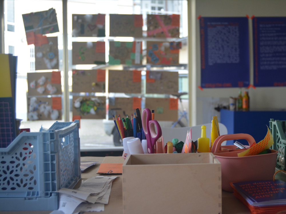 Das Foto zeigt den Basteltisch, an dem die Teilnehmer*innen ihre Stadtmodelle bauen konnten. Verschiedene Boxen (in die man nicht schauen kann) enthalten Materialien, eine große Stiftebox enthält etwa 30 Stifte, eine Schere und Flaschen mit Kleberr.