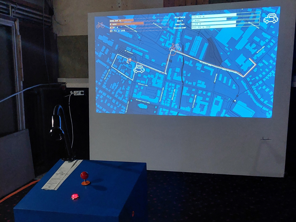 Das Foto zeigt einen hell erleuchteten Bildschirm in einem dunklen ehemaligen Kinosaal. Der Bildschirm zeigt eine Straßenkarte, getaucht in blau. Zwei Wege sind eingezeichnet, die zum einen den Weg mit dem Auto und zum anderen den Weg mit dem Fahrrad zeigen. Vorne links im Bild ist eine blaue Box mit einem roten Joystick zu sehen.