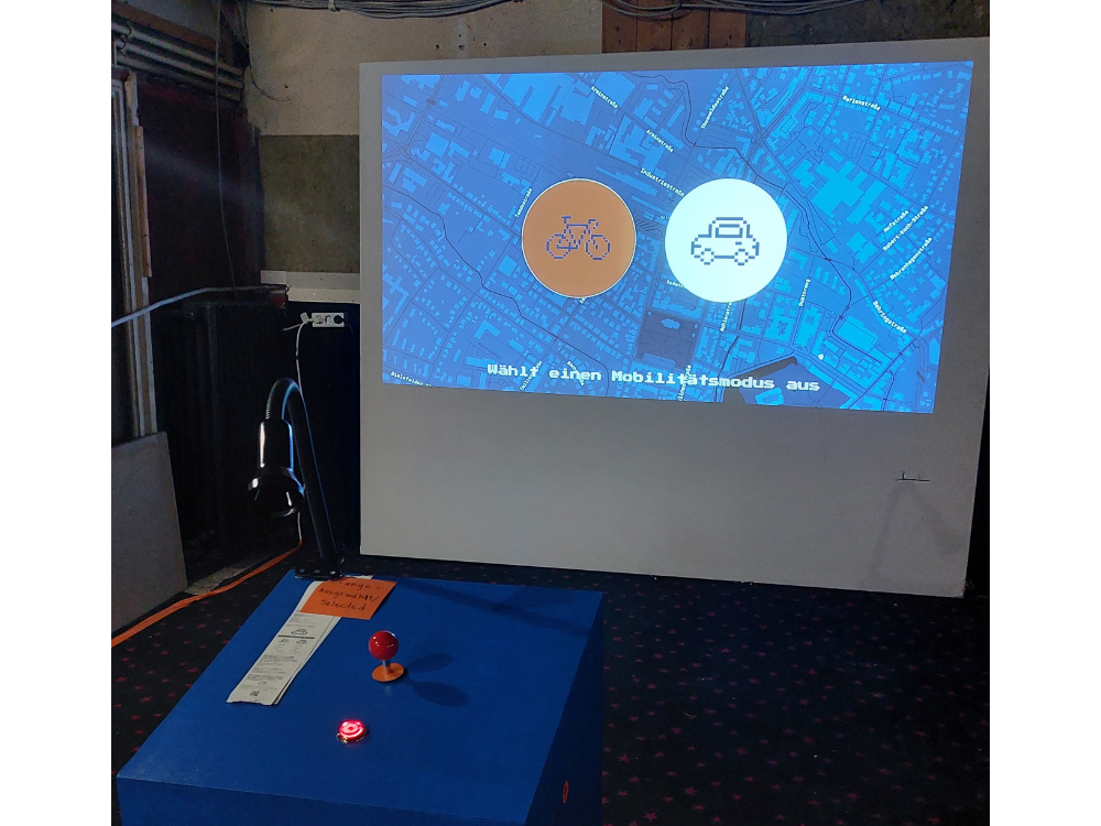 Das Foto zeigt einen hell erleuchteten Bildschirm in einem dunklen ehemaligen Kinosaal. Der Bildschirm zeigt eine Straßenkarte, getaucht in blau, präsent vorne links ein orangener Kreis mit einem blauen Fahrrad, rechts daneben ein weißer Kreis mit einem Auto. Vorne links ist außerdem noch eine blaue Box mit einem roten Joystick zu sehen.