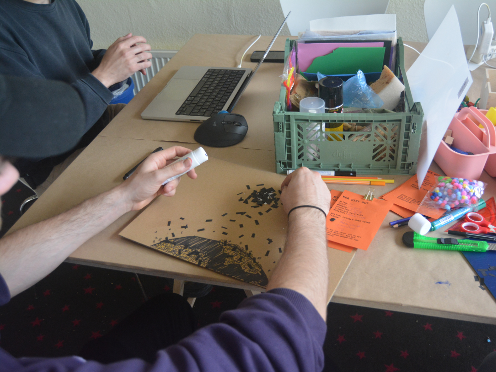 An einem Tisch sitzen zwei Personen über Eck, von denen jeweils nur die Hände zu sehen sind. Die Person im Vordergrund arbeitet an ihrem Stadtmodell und klebt Papierschnipsel auf, während die Person im Hintergrund an einer Laptoptastatur sitzt. Eine grüne Plastikbox auf dem Tisch beinhaltet Bastelmaterial.