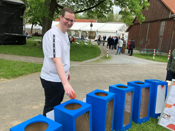 Das Foto zeigt sechs blaue Abstimmungssäulen für Bälle im Vordergrund, die sich in einem Halbkreis durchs Bild ziehen. Hinter den Säulen steht ein junger Mann mit Brille. Er lächelt und wirft gerade einen roten Ball in die zweite Säule von links. 