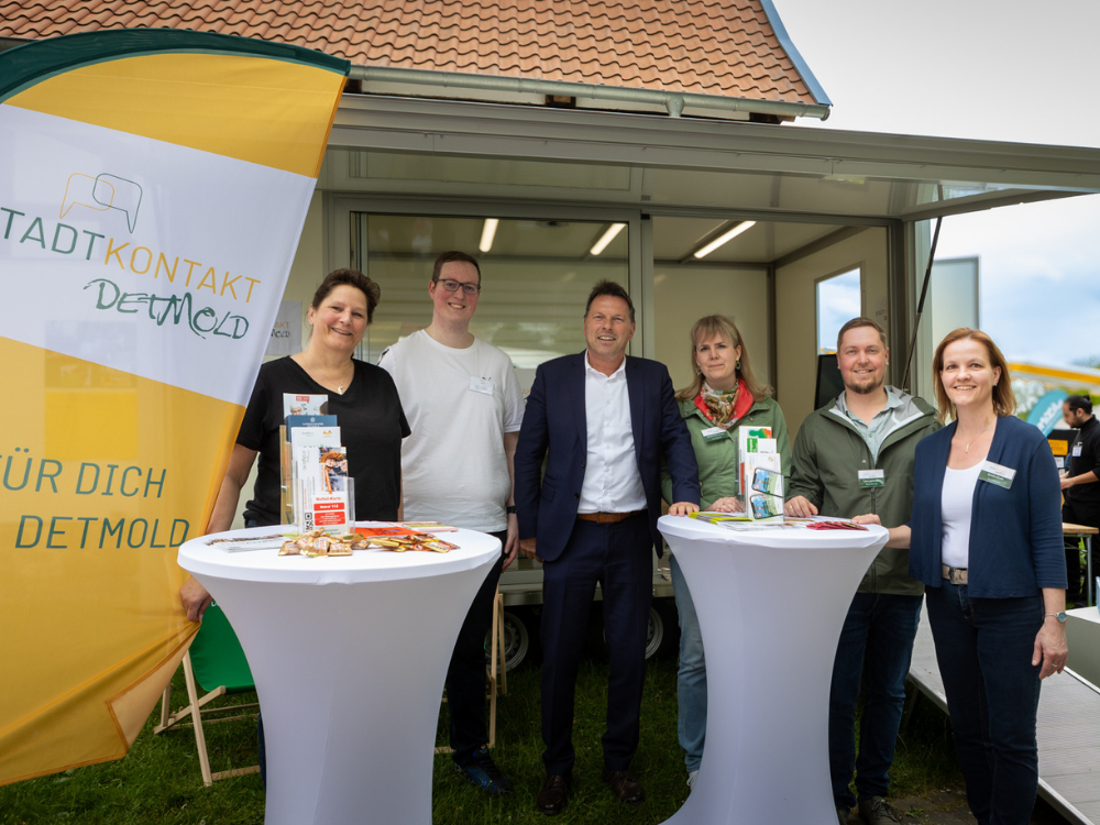 Auf dem Foto stehen sechs Mitarbeiter*innen der Stadt Detmold nebeneinander und lächeln in die Kamera. Links ragt eine Beachflag in weiß-orange, mit dem Aufdruck "Stadtkontakt Detmold. Für Dich. Für Detmold." ins Bild.