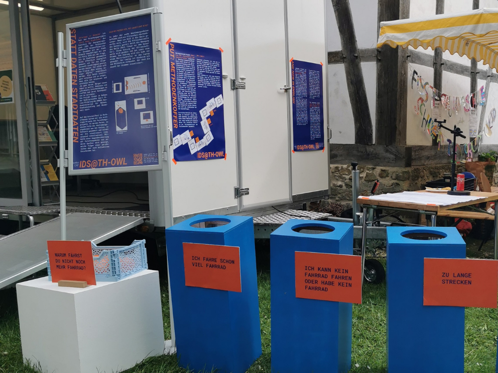 Das Foto zeigt drei blaue Abstimmungssäulen für Bälle im Vordergrund. Im Hintergrund sieht man angeschnitten den weißen Anhänger der Stadt Detmold sowie eine große Wand mit weiterem Infomaterial. Die Schrift ist so klein, dass man auf dem Foto nichts lesen kann.