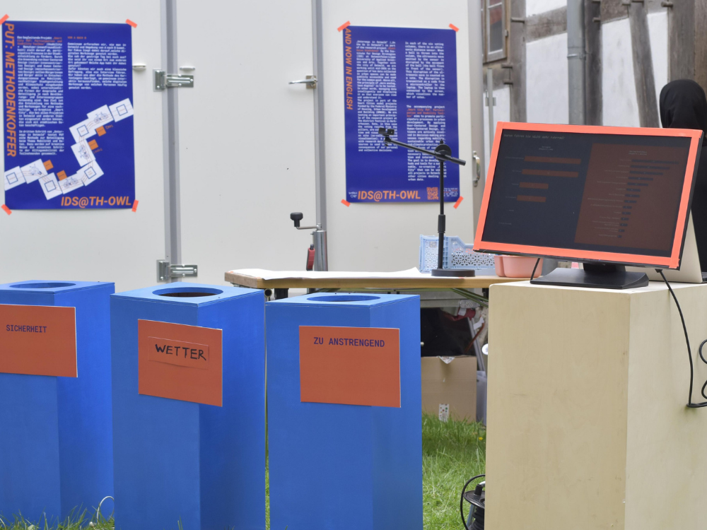 Das Foto zeigt drei blaue Abstimmungssäulen für Bälle im Vordergrund. Im Hintergrund sieht man angeschnitten den weißen Anhänger der Stadt Detmold sowie zwei große Infowände mit weiterem gedrucktem Infomaterial. Die Schrift ist so klein, dass man auf dem Foto nichts lesen kann. Rechts davon steht ein großer Monitor mit orangenem Rahmen auf einer hohen viereckigen Säule.