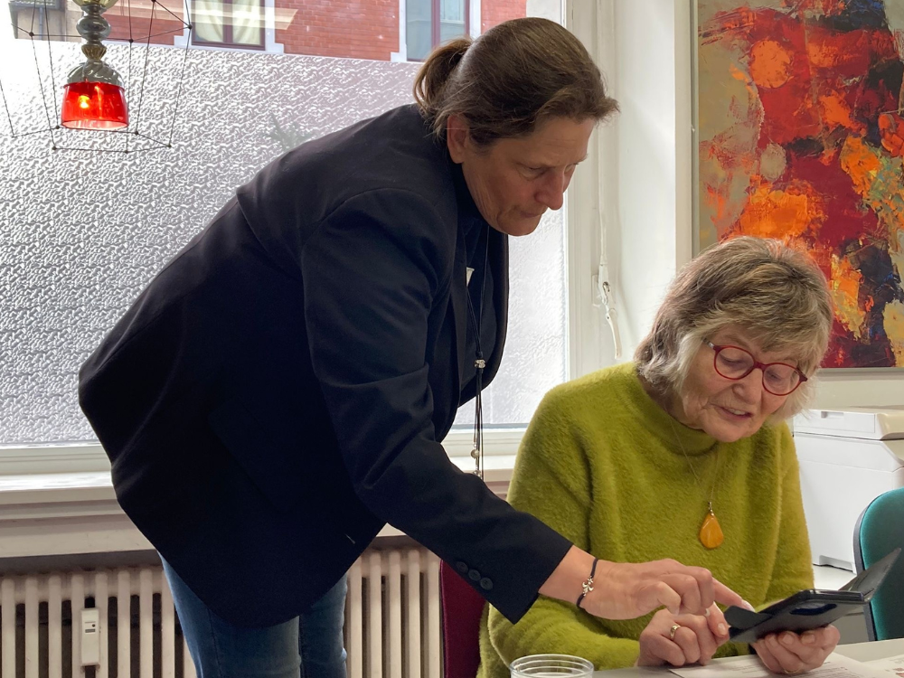 Eine Frau im schwarzen Blazer erklärt einer Seniorin an ihrem Smartphone das Scannen von QR-Codes.