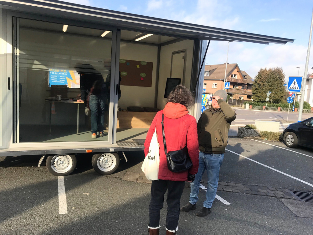 Der Anhänger steht aufgeklappt auf dem Supermarktplatz. Davor unterhält sich ein Mitarbeiter der Stadt mit einer Bürgerin. Die Sonne scheint. 