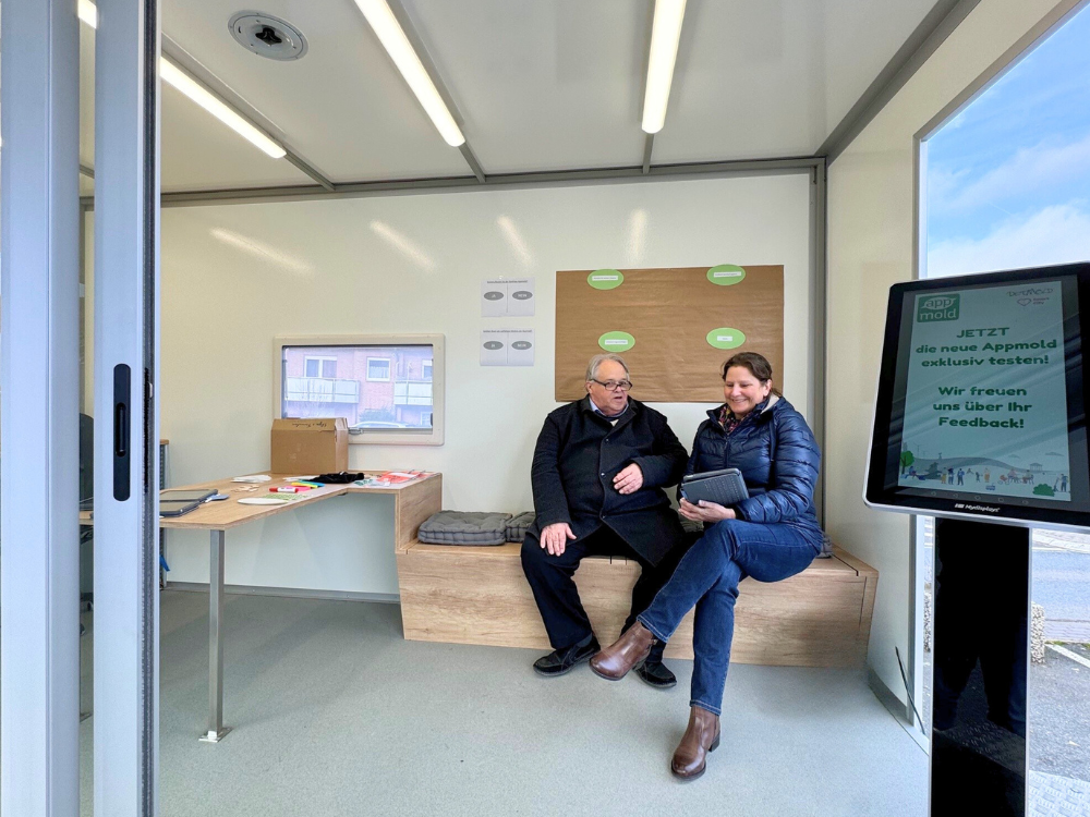 Das Foto zeigt eine Mitaerbeiterin der Stadt Detmold im Gespräch mit dem Ortsteilbürgermeister. Beide tragen warme Jacken und tauschen sich über Informationen auf einem Tablet aus, das die Frau in der Hand hält. Beide sitzen auf einer Holzbank.