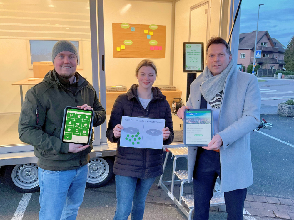 Das Foto zeigt zwei Mitarbeitende der Stadt Detmold und den Bürgermeister Frank Hilker, die Infomaterial in die Kamera halten. Im Hintergrund sieht man die große Glaswand des städtischen Anhängers.