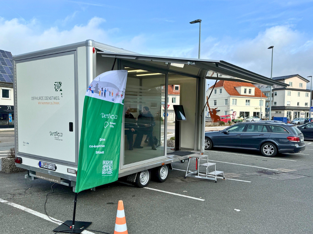 Der Anhänger steht aufgeklappt auf dem Supermarktplatz. Eine Beachflag weist auf den Aktionstag hin. Im Hintergrund parken Autos.