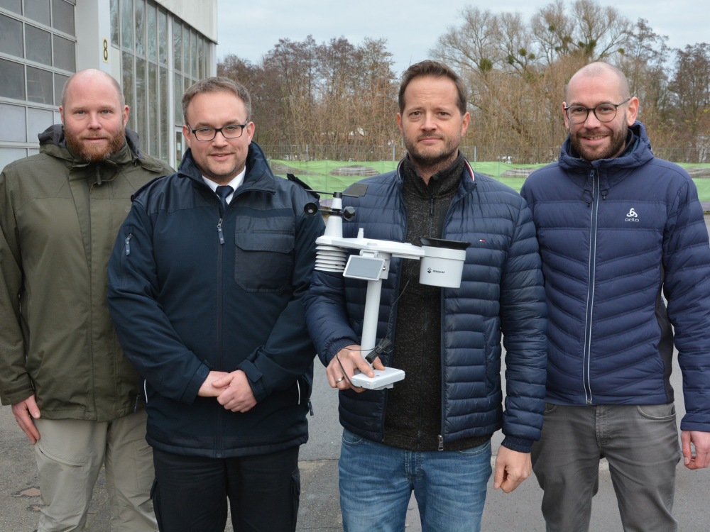 Vier Herren stehen nebeneinander in dicken Jacken vor einer Garage und halten eine Wetterstation in die Kamera.
