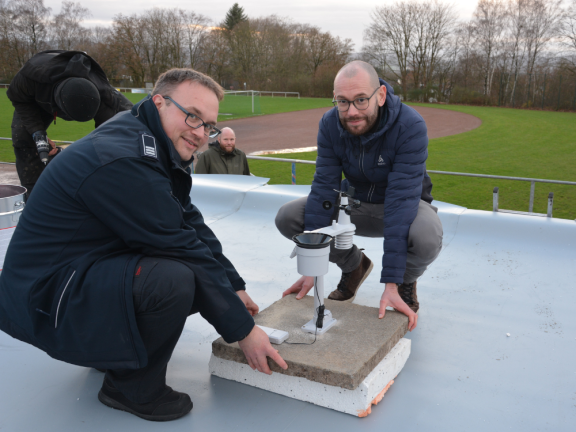 Zwei Männer hocken auf dem Dach des Sporthauses und installieren die erste Wetterstation, die auf einer Betonplatte befestigt ist. Unter der Platte befindet sich noch eine dicke weiße Platte. 
