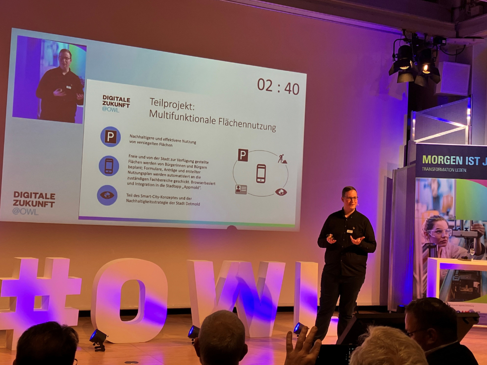 Das Bild zeigt die Bühne, dort hält ein junger Mann mit Brille eine Präsentation. Auf der Bühne stehen weiße Modelle der Buchstaben #OWL.