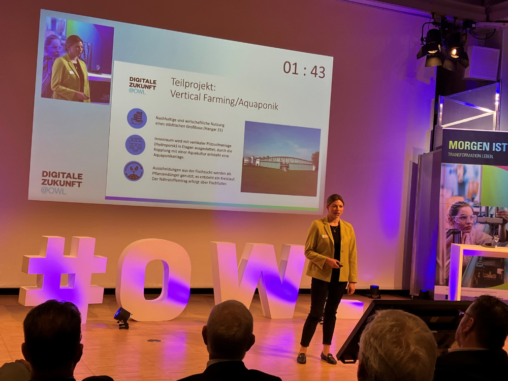 Das Bild zeigt die Bühne, dort hält eine junge Frau mit Zopf eine Präsentation. Auf der Bühne stehen weiße Modelle der Buchstaben #OWL.
