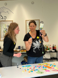 Zwei Personen bei einem Workshop, eine hält eine mit bunten Kugeln gefüllte Flasche, auf dem Tisch liegen bunte Plastikdeckel und Zettel mit Notizen.