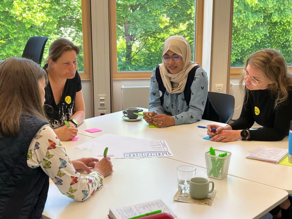 Vier Frauen sitzen an einem Tisch und arbeiten gemeinsam an einem Workshop mit Papier und Stiften, im Hintergrund grüne Bäume durch Fenster sichtbar. 