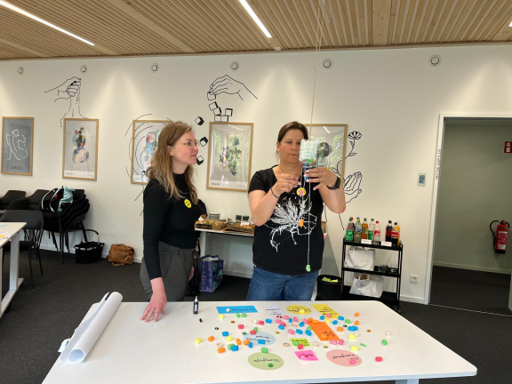 Zwei Personen stehen an einem Tisch mit bunten Würfeln und Zetteln, eine hält eine abgeschnittene Plastikflasche. Daraus wird die Lösung für den digitalen Methodenkoffer visualisiert 