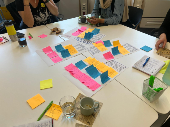 Workshop-Tisch mit mehreren Personen, auf dem bunte Haftnotizen auf ausgedruckten Arbeitsblättern verteilt sind, daneben Notizbücher, Stifte, Getränke und Kekse 