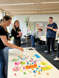 Mehrere Personen in einem Workshopraum verteilen bunte Papierstücke auf einem Tisch mit beschrifteten Karten und Notizen an der Wand 