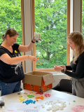 Zwei Frauen stehen an einem Tisch mit bunten Perlen und einem Karton, eine hält eine weiße Kette mit aufgefädelten bunten Styroporchips hoch, die andere hält eine Schnur mit Perlen.