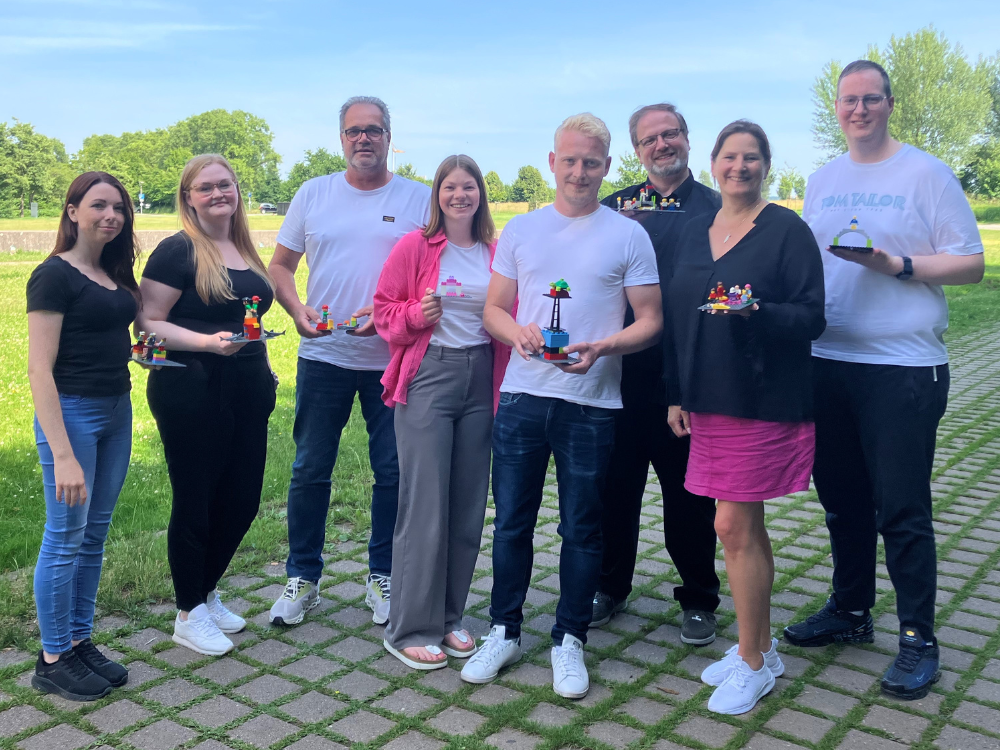 Acht Teammitglieder stehen im Freien auf gepflastertem Weg, halten ihre bunten LEGO-Modelle und lächeln. 