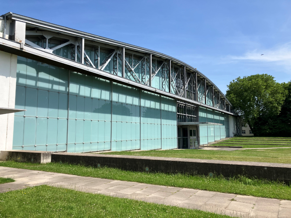 Bild zeigt die Eventlocation „Hangar 21" mit großer Glasfassade und sichtbarer Stahlkonstruktion unter blauem Himmel, umgeben von Rasenflächen und Bäumen.