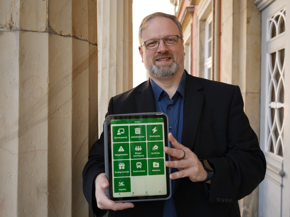 Dennis Schäffer, CDO der Stadt Detmold, steht vor dem Rathaus und hält ein iPad mit der Stadtapp "Appmold" bildfüllend in die Kamera.