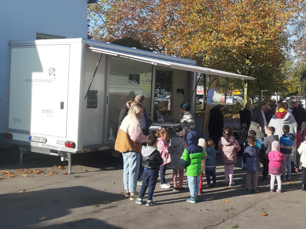 Der städtische Anhänger parkt auf einem weitläufigen Parkplatz. Aktuell besucht ein Kindergarten mit vier Mitarbeiter*innen und vielen Kindern den Stadtkontakt mobil. Alle stehen mit dem Rücken zur Kamera.