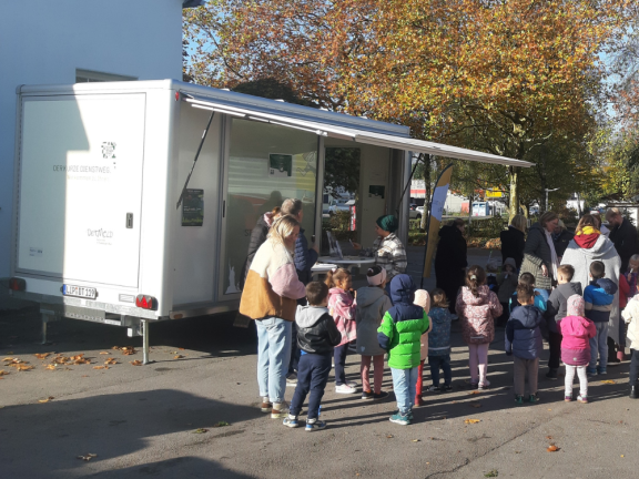 Der städtische Anhänger parkt auf einem weitläufigen Parkplatz. Aktuell besucht ein Kindergarten mit vier Mitarbeiter*innen und vielen Kindern den Stadtkontakt mobil. Alle stehen mit dem Rücken zur Kamera. 