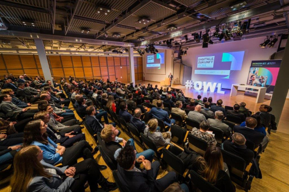 Das Bild zeigt den Vortragssaal in der Totale. Man sieht geschätzt 100 Personen die auf aufsteigenden Sitzreihen. Platz genommen haben. im Hintergrund sieht man die große Bühne, auf der aktuell nur die großen weßen Buchstaben 