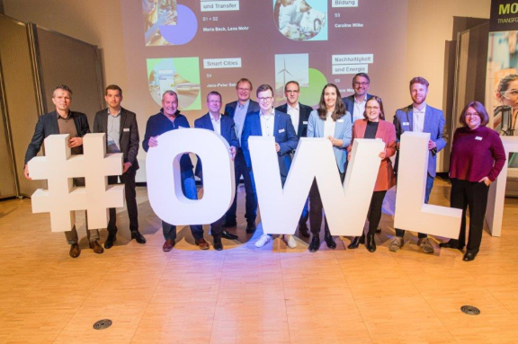 Das Bild zeigt die Bühne mit ca. 15 Menschen. Diese halten große weiße Buchstaben, die das Wort 