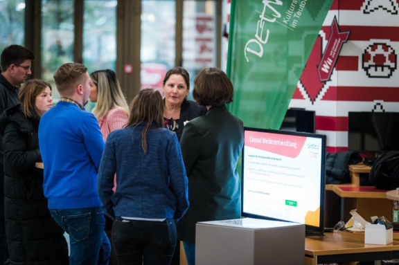 Man sieht eine Mitarbeiterin der Stadt Detmold am Stand, die mit einer Dame (nur von hinten zu sehen) im intensiven Gespräch ist. Weitere Personen hören interessiert zu. 