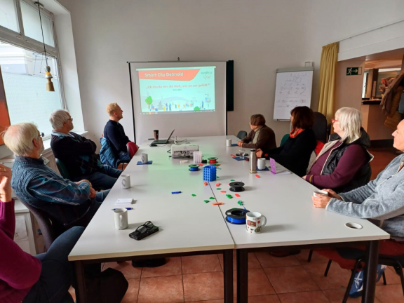 Das Foto zeigt einen großen weißen Tisch in einem abgedunkelten Raum. Man sieht sieben ältere Personen und einen jüngeren, blonden Mann, der durch die Präsentation führt. 