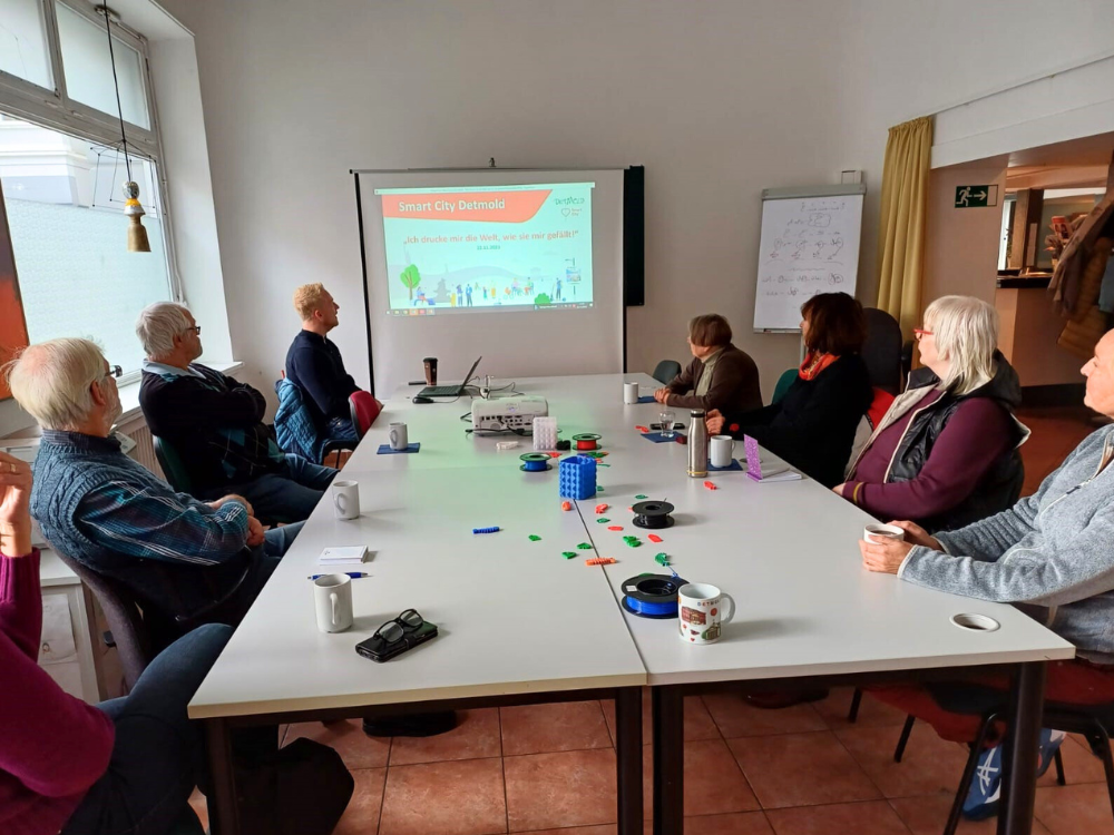 Das Foto zeigt einen großen weißen Tisch in einem abgedunkelten Raum. Man sieht sieben ältere Personen und einen jüngeren, blonden Mann, der durch die Präsentation führt.